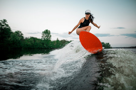 Young attractive brunette girl jumping on the orange wakesurf on high wave of motorboat on the...