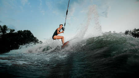 Active brunette woman riding on the wakesurf holding a rope of a motorboat on the background of treesの写真素材
