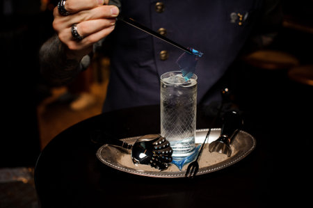Bartender with tattoo decorating fresh and sweet summer cocktail with blue piece of caramel on the...
