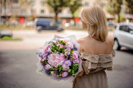 Rear view blonde girl in beige dress holding in her hands a bouquet of tender pink and violet flowers decorated with green leavesの写真素材