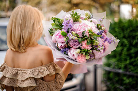 Rear view blonde woman in beige dress holding in her hands a bouquet of tender pink and violet flowers decorated with green leavesの写真素材