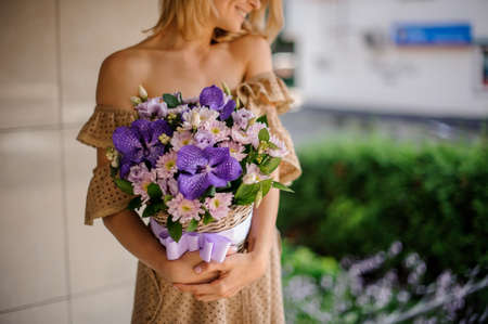 Young smiling girl in beige dress holding in her hands a basket of pink chrisantemum and violet orchids decorated with green leavesの写真素材