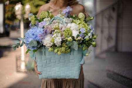 Young beautiful girl in beige dress holding in her hands a huge blue basket of tender white and blue flowers decorated with green leavesの写真素材