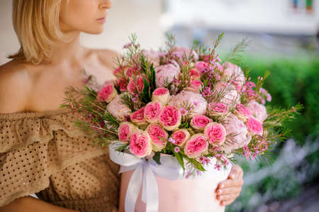Young girl in beige dress holding in her hands a box of tender pink roses and peonies decorated with green branchesの写真素材