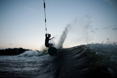 Young wakesurfer dressed in swimsuit riding on the blue river waves with splashes on summer evening