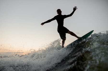 Dark silhouette of a young man riding on a wake board at the summer evening