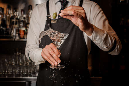 Barman holding a splashing fresh and strong summer martini cocktail with a green olive against the barの写真素材