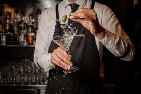 Bartender holding a splashing fresh and strong summer martini cocktail and a green olive against the barの写真素材
