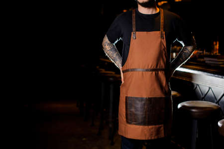 Bartender with tattoo on hands dressed in brown apron standing near the dark bar counterの写真素材