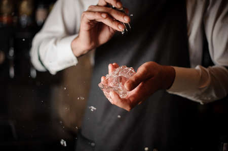 Professional male bartender breaking an ice cube to prepare a fresh cocktail with a special tool at the bar counterの写真素材