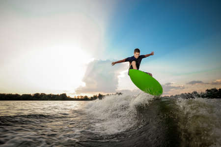 Young active brunet man riding on the green wakeboard on high wave of motorboat, having healthy summertimeの写真素材