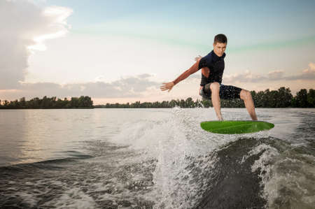 Young active brunet man jumping on the green wakeboard on high wave of motorboat, having healthy summertimeの写真素材