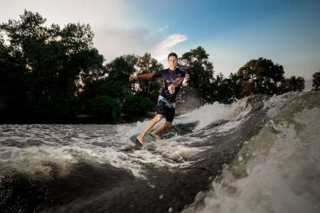 Young attractive man riding on the wakeboard on high wave of motorboat on the background of treesの写真素材