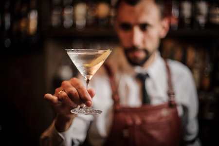Bartender in the white shirt and brown leather apron holding a transparent cocktail in the martini...
