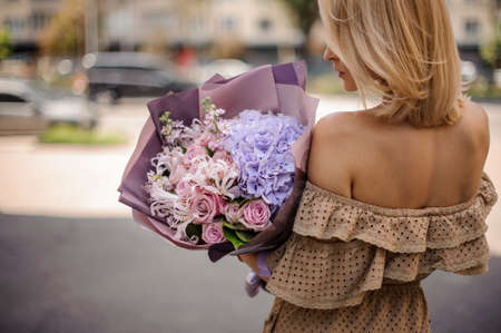Back view of blonde young woman in beige dress holding a romantic bouquet of flowers in purple tonesの写真素材