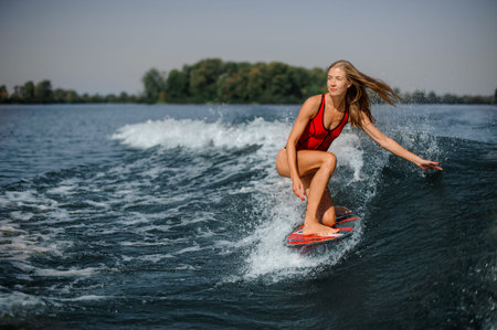 Attractive slim blonde woman wakesurfer riding down the blue splashing wave on a warm dayの写真素材
