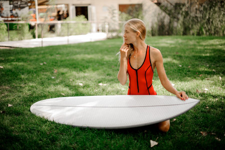 Beautiful and young blonde woman dressed in red swimsuit sitting on green lawn with a wakeboard on...