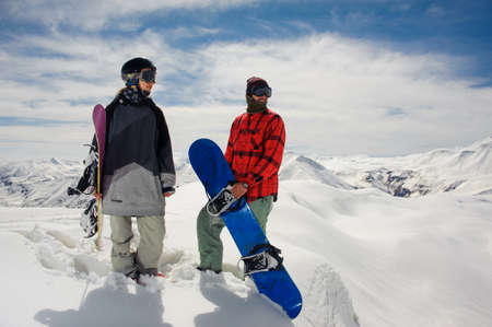 guy and a girl in warm clothes and ski goggles stand with snowboards in their hands against the blue sky and mountain snow topsの写真素材