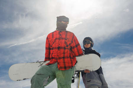 guy and a girl in warm clothes and ski goggles stand on the snow with snowboards in their hands against the blue skyの写真素材