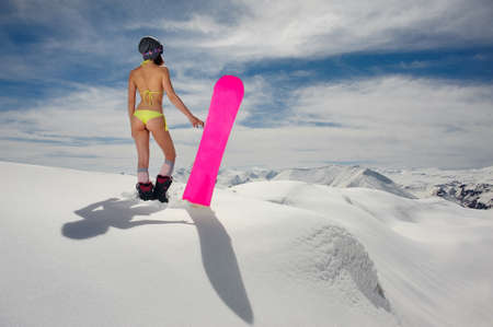 Rear view of young sexy girl dressed in a bright swimsuit standing on the slope with a snowboard in the handsの写真素材