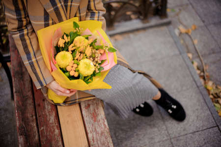 Top view of woman in long dress and plaid coat sitting on bench with a bouquet of yellow flowersの写真素材