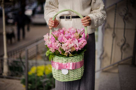 Woman in white sweater holding a beautiful green wicker basket with pink flowers against the cityの写真素材