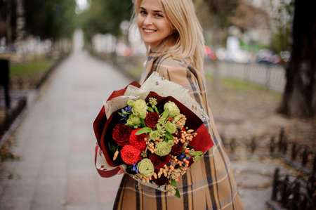 Happy smiling blonde woman in plaid coat holding a bright bouquet of flowers walking on alley in the autumn parkの写真素材