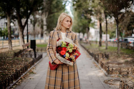 Blonde woman in plaid coat holding a bright bouquet of flowers standing on alley in the parkの写真素材