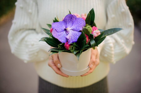 Girl in the white sweather holding in her hands a white box of violet orchid decorated with little...