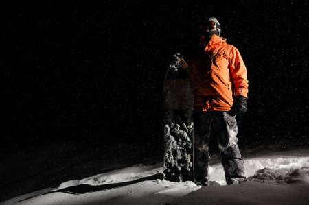 Snowboarder in orange sportswear and mask standing with a board on a snowy hill at nightの写真素材