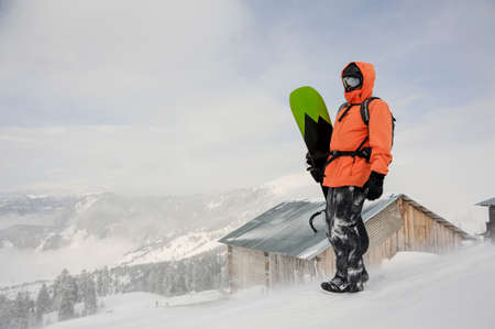 Man standing with the snowboard in hands onthe hill in the mountain in the popular tourist resort in Goderzi, Georgia on the background of snowcovered houseの写真素材