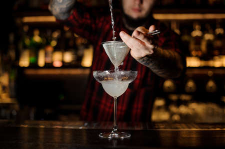 Barman pouring fresh Margarita cocktail into a cocktail glass using a strainer on the bar counterの写真素材