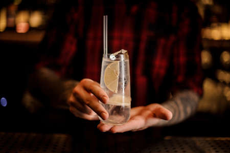 Man holding elegant long drink glass filled with Tom Collins cocktail against the lights of barの写真素材