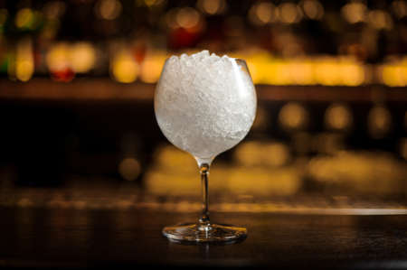 Gin Tonic cocktail glass full of crushed ice standing on the steel bar counter on the blurred dark and light backgroundの写真素材