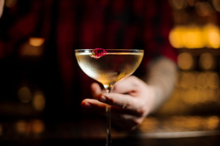 Bartender serving a glass of a cocktail Twinkle withone berry on the bar counter on the blurred dark...