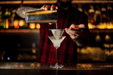 Bartender pourring a White Lady cocktail from the steel shaker through the sieve to a glass on the...
