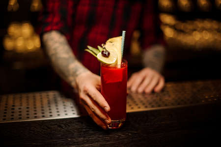 Bartender with tattoos holding a glass of red sweet juicy drink in a cocktail glass with fruitの写真素材