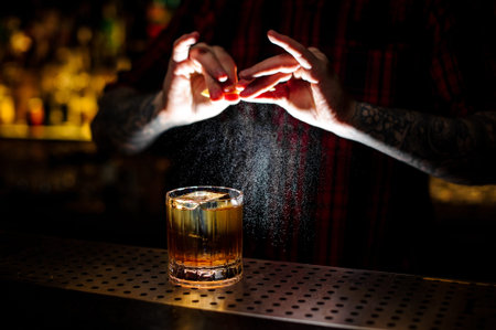 Barman splashing orange peel juice into a strong cocktail in the dark background of bar
