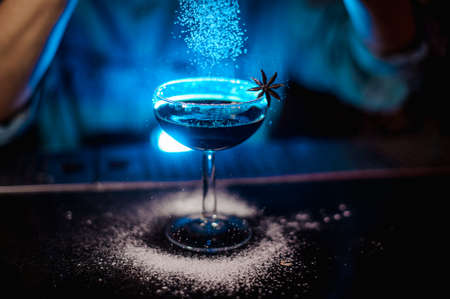 Girl adding to a cocktail decorated with badian a sugar powder through the strainer in the blue lightの写真素材