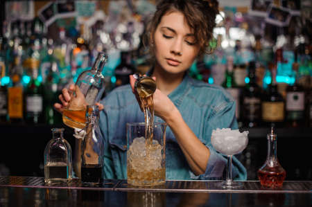 Bartender girl pouring to the measuring glass cup with ice cubes an alcoholic drink from jiggerの写真素材