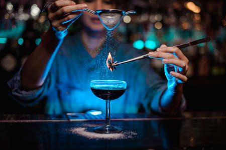 Girl adding to a brown cocktail and pour on a flamed badian on tweezers a powdered sugar in the blue lightの写真素材
