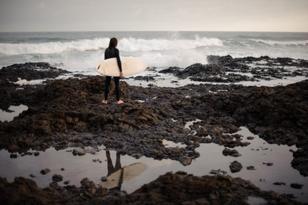 Rear view brunette man standing with a surf in his hands on the rock beach looking at the ocean.