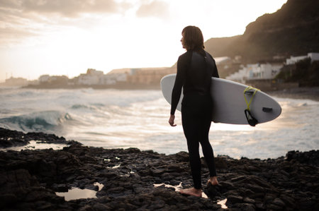Rear view brunette boy standing with a surf in his hands on the rock beach looking at the sea on the...