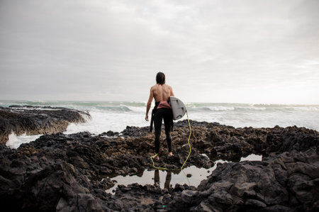 Rear view brunette boy standing in the black swimsuit with naked torso with a white surf on the rock...
