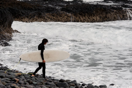 Brunette boy in the black swimsuit with a white surf in his hands on the pebble beach walking on the...