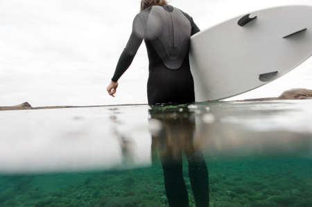 Strong brunette guy standing in the black swimsuit with a white surf in his hands in the ocean water.の写真素材