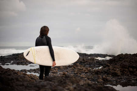 Rear view brunette man standing in the black swimsuit with a surf in his hands on the shoreの写真素材