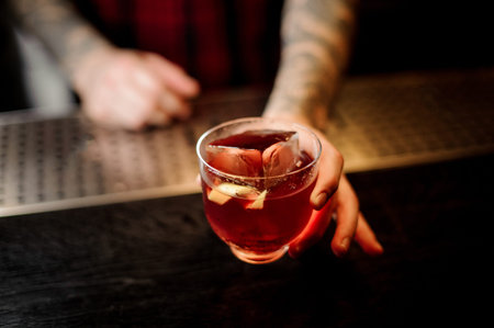 Bartender tattooed hand holding a strong alcoholic drink with whiskey and orange peel