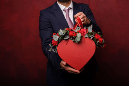 Man holding in hands a red box of beautiful flowers standing on the background of the dark wallの写真素材