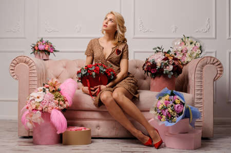Beautiful girl in the beige dress sitting on the couch surrounded by the boxes and bouquets of flowers on the background of white wallの写真素材